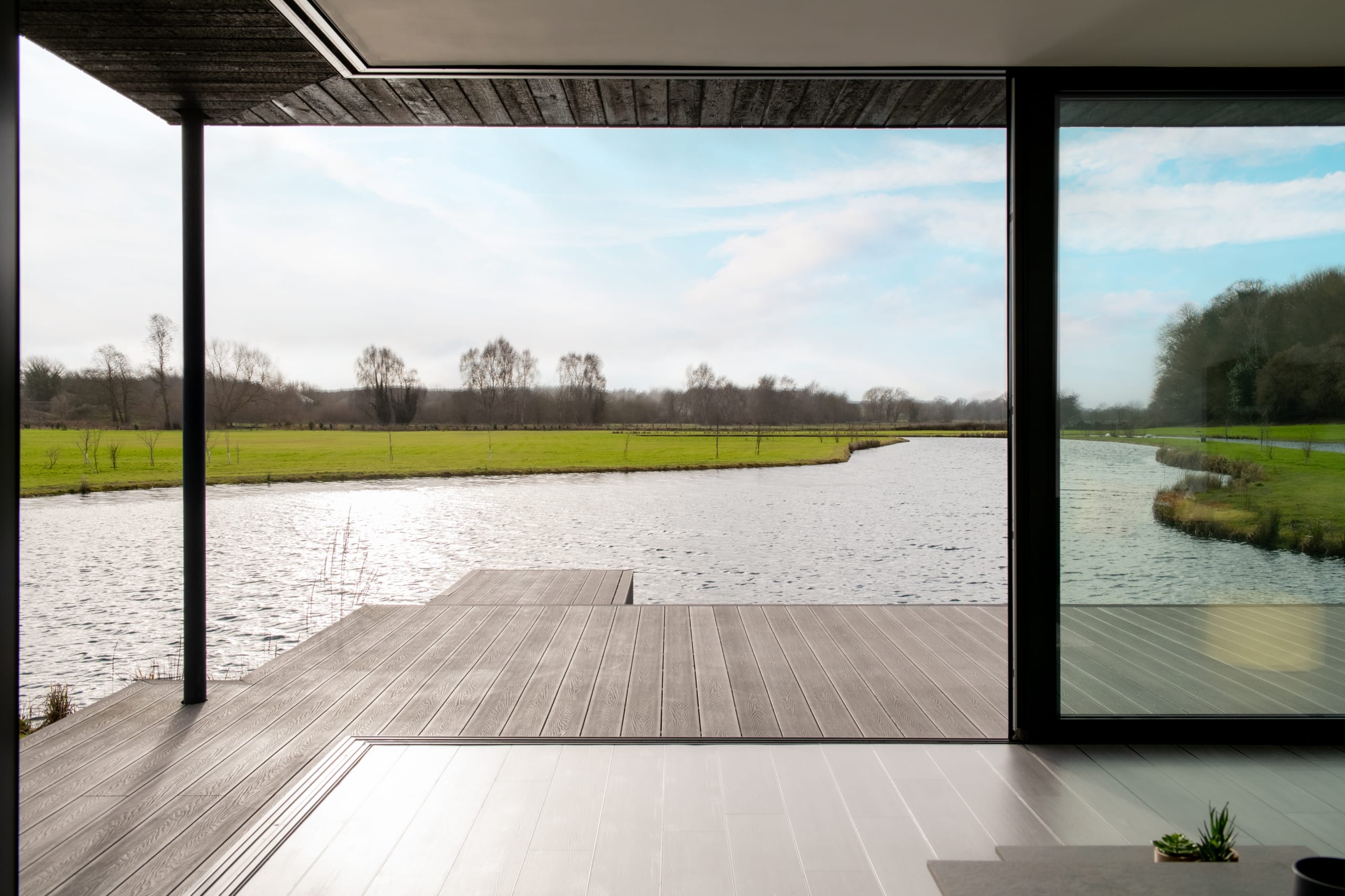 Modern interior opening to wooden deck overlooking serene river and green countryside through floor-to-ceiling glass walls